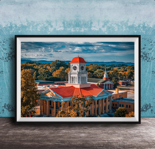 A framed photograph of a historic building with a clock tower, surrounded by trees and mountains in the background.