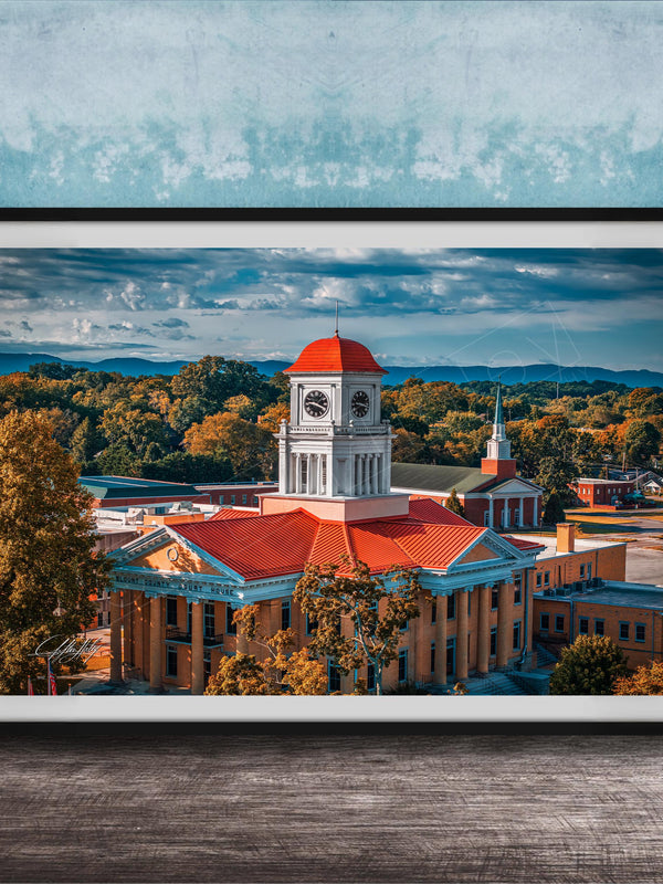 A framed photograph of a historic building with a clock tower, surrounded by trees and mountains in the background.