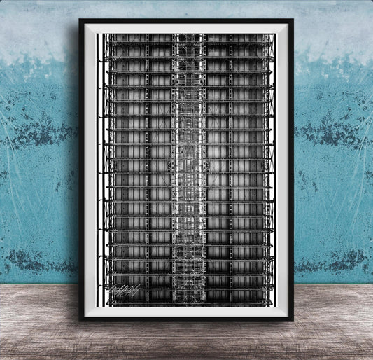 The image is a black and white photograph of a tall, narrow building with a grid-like pattern of windows and steel beams. The photograph is framed and displayed on a wooden surface against a blue wall.