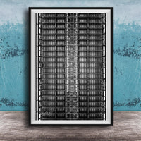 The image is a black and white photograph of a tall, narrow building with a grid-like pattern of windows and steel beams. The photograph is framed and displayed on a wooden surface against a blue wall.