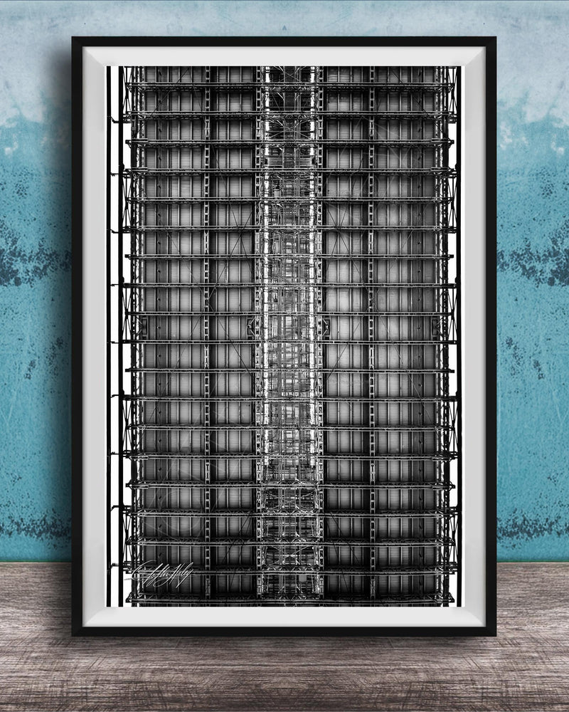 The image is a black and white photograph of a tall, narrow building with a grid-like pattern of windows and steel beams. The photograph is framed and displayed on a wooden surface against a blue wall.