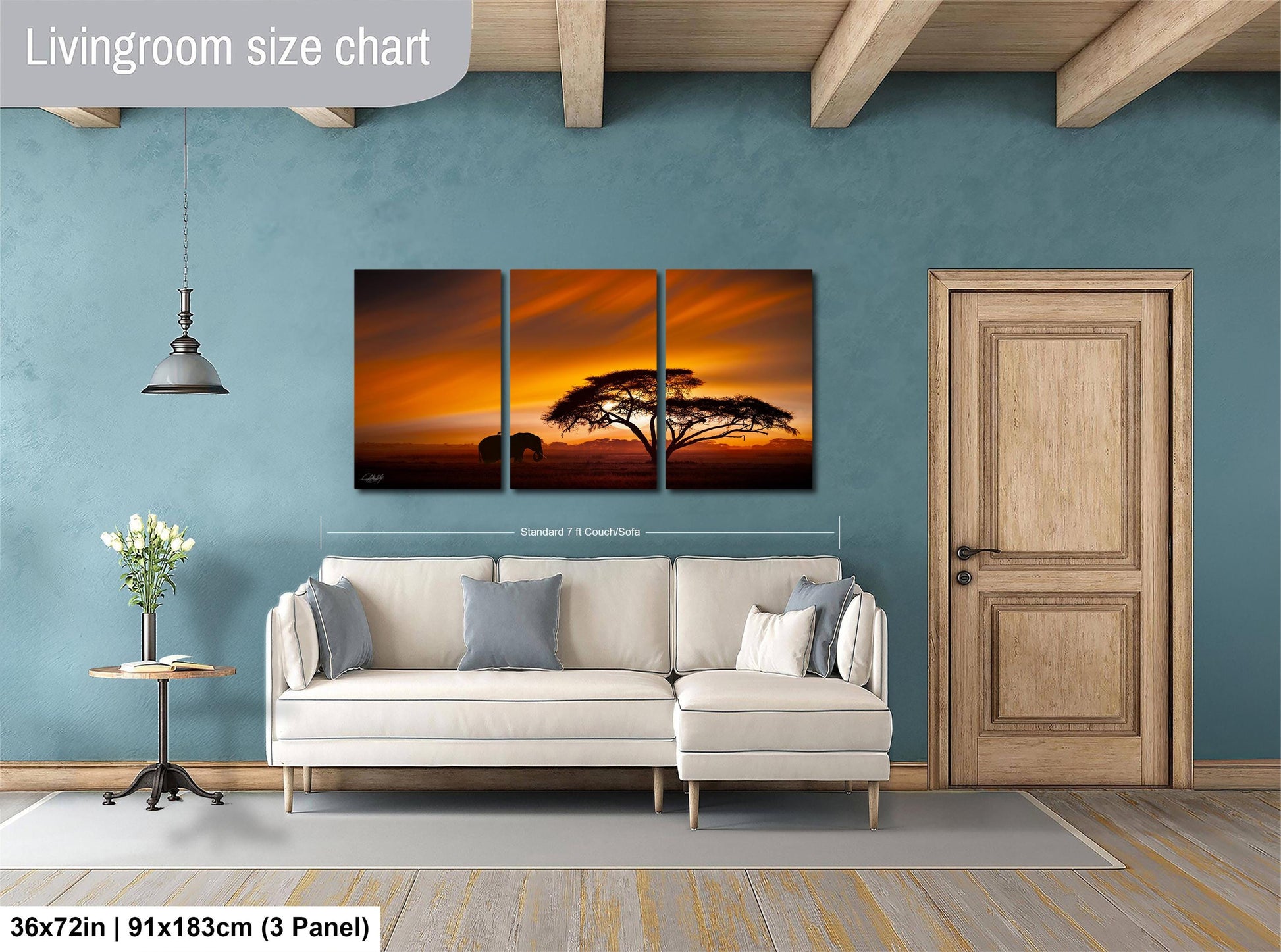 The image is a living room with a large wall art depicting a sunset over a savanna landscape. The wall art is divided into three panels, with the largest panel spanning approximately 36 inches by 21 inches. The smaller panels are smaller in size, with one panel measuring approximately 11.85 inches by 3.25 inches and another measuring approximately 11.85 inches by 3.25 inches. The room also includes a white sofa, a wooden door, and a small table with a vase of flowers.