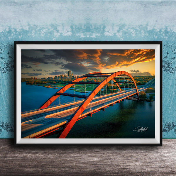 The image is a framed photograph of a red arch bridge spanning across a body of water, with a city skyline visible in the background.