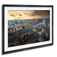 The image is a framed aerial view of a cityscape, featuring a mix of tall buildings, roads, and a river. The sky is filled with clouds, creating a dramatic backdrop for the city's skyline. The image is framed in a black frame and is hung on a wall.