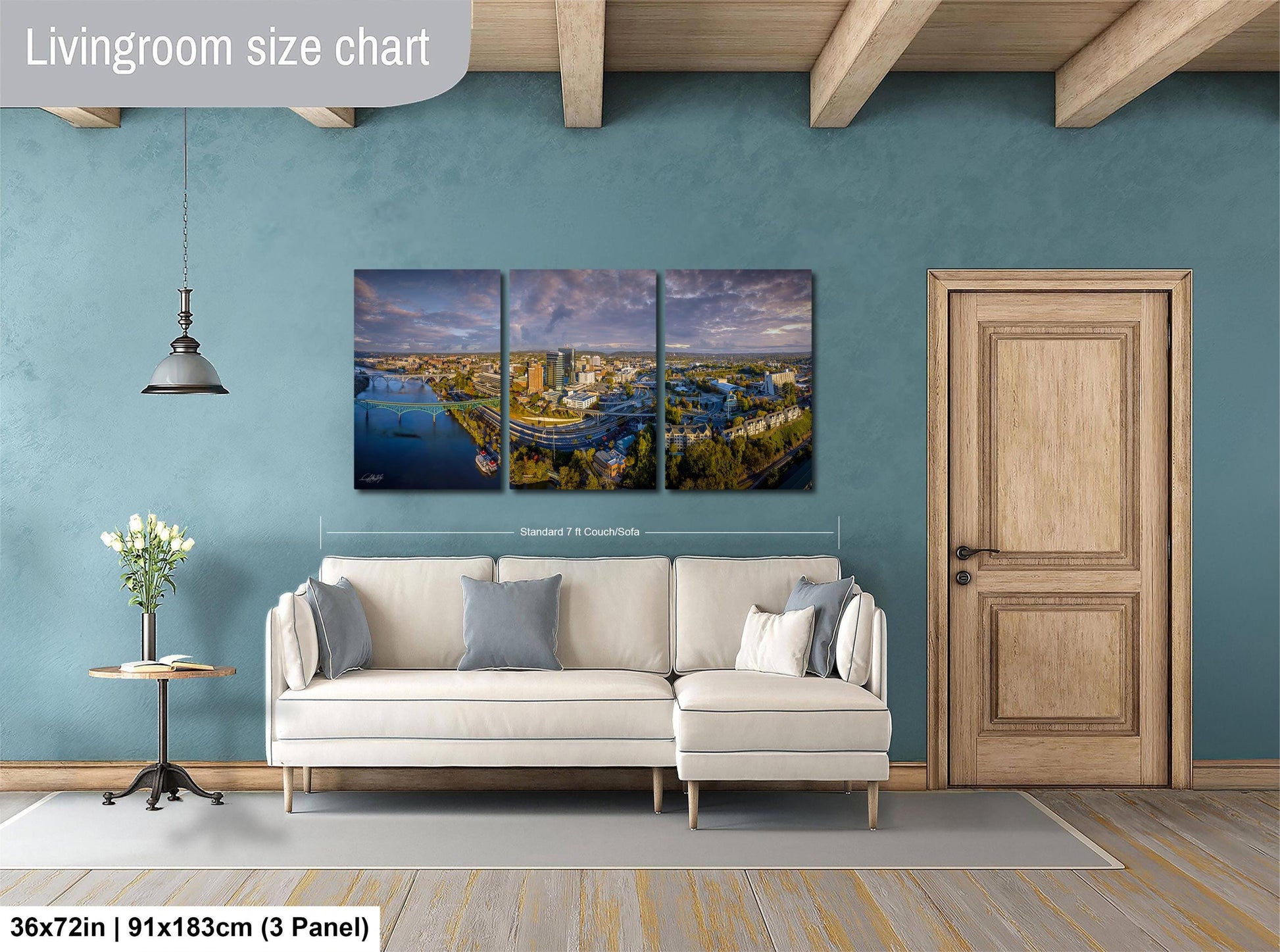 The image is a living room with a large, three-panel landscape painting hanging on the wall. The painting depicts a cityscape with a river and buildings. The room has a white sofa, a wooden door, and a small table with a vase of flowers. The painting is approximately 36 inches wide and 15 inches tall, and it is mounted on a wooden beam ceiling.