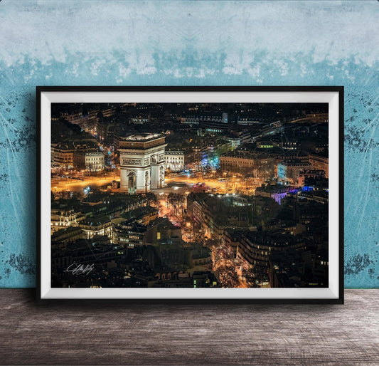 The image is a framed photograph of a city at night, featuring a large, illuminated archway or monument in the center. The cityscape is filled with numerous buildings and lights, creating a vibrant and bustling nighttime scene.