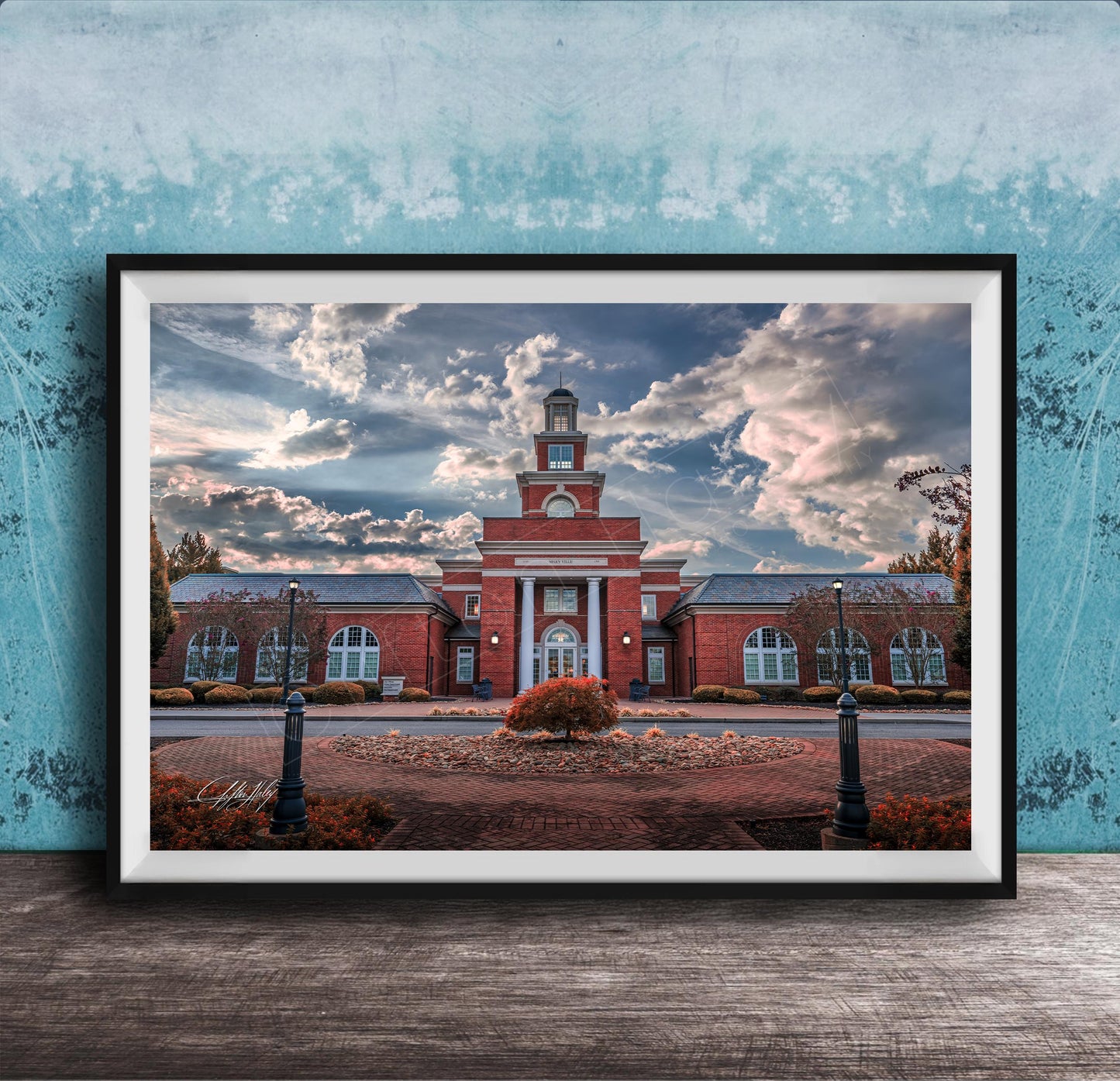 A framed photograph of a red brick building with a clock tower, surrounded by a brick walkway and landscaping, set against a cloudy sky.