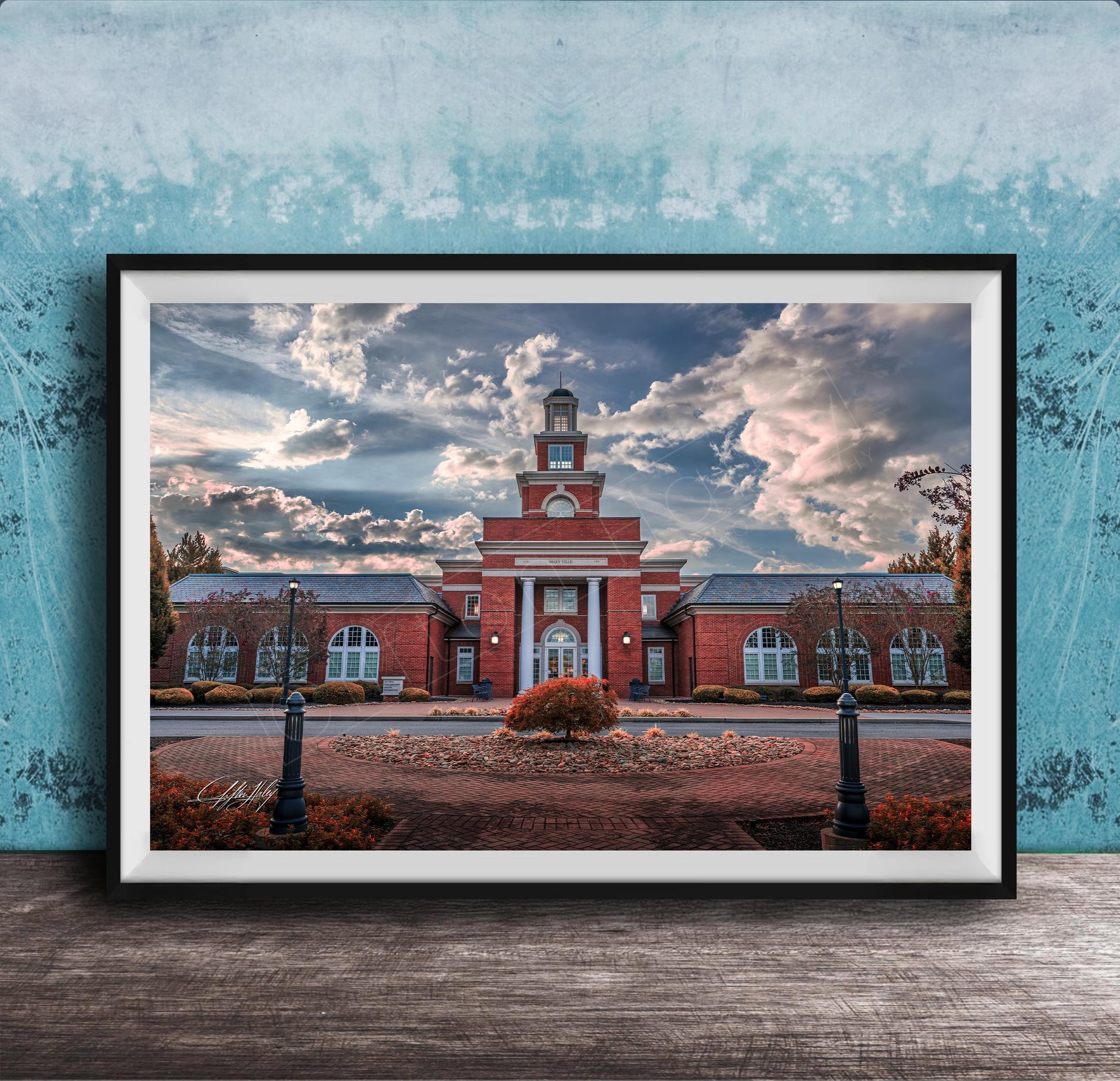 A framed photograph of a red brick building with a clock tower, surrounded by a brick walkway and landscaping, set against a cloudy sky.