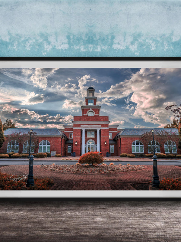 A framed photograph of a red brick building with a clock tower, surrounded by a brick walkway and landscaping, set against a cloudy sky.