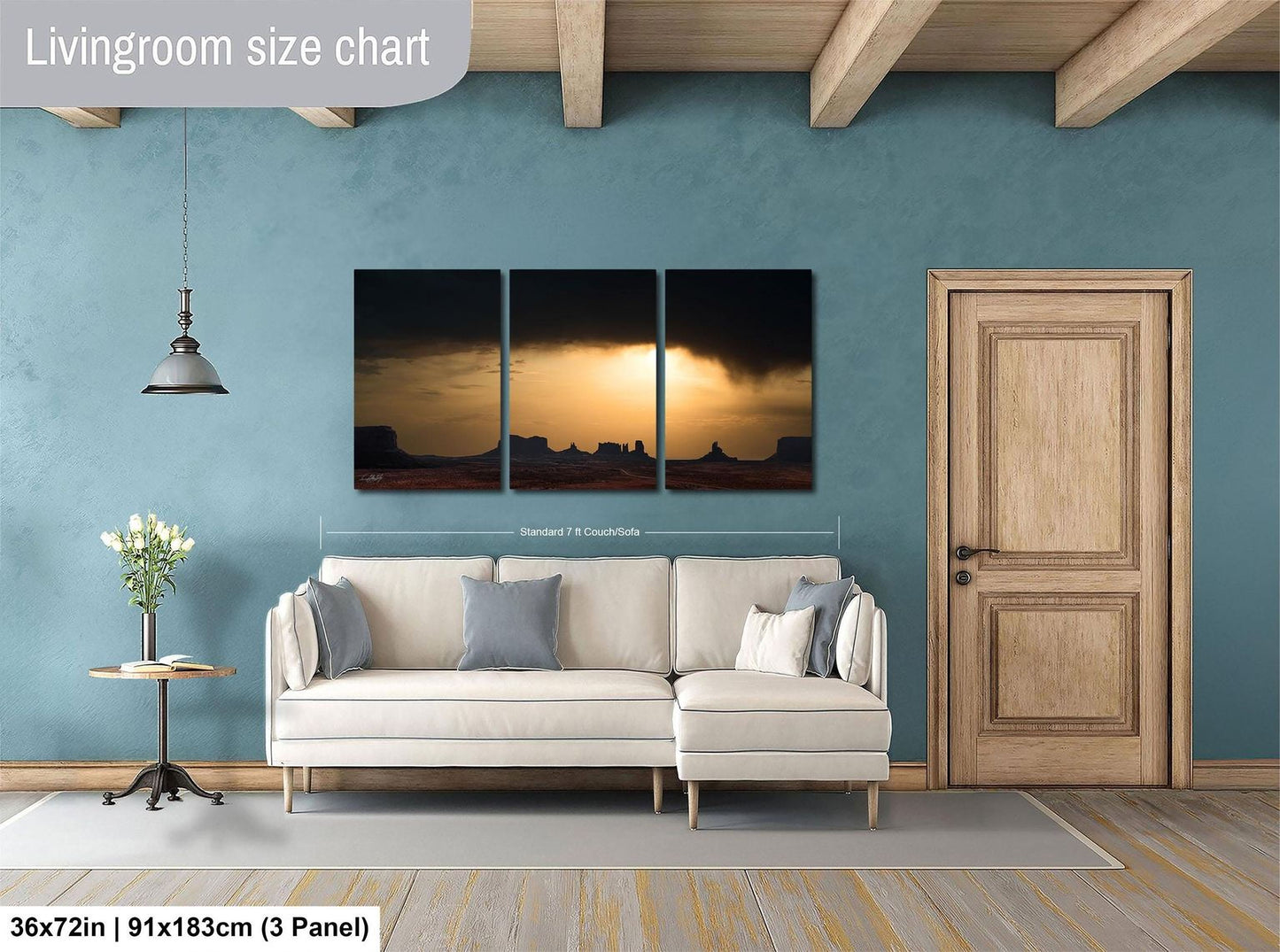 The image is a living room with a large wall art piece depicting a desert landscape with a sunset. The living room has a white sofa, a wooden door, and a wooden table with a vase of flowers. The wall art is approximately 36 inches wide and measures approximately 91 inches tall.