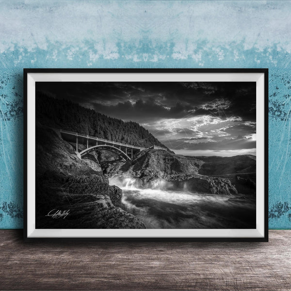 A black and white photograph of a bridge spanning over a rushing river, with dramatic clouds in the sky above.