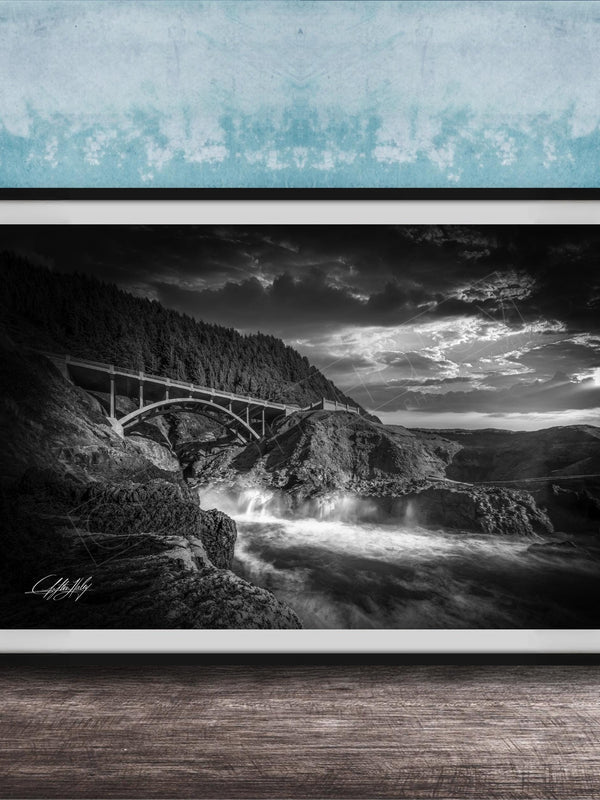 A black and white photograph of a bridge spanning over a rushing river, with dramatic clouds in the sky above.