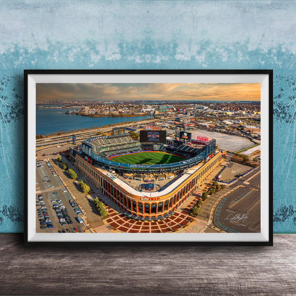 An aerial view of a baseball stadium with a large field, surrounding parking lots, and a body of water in the background.