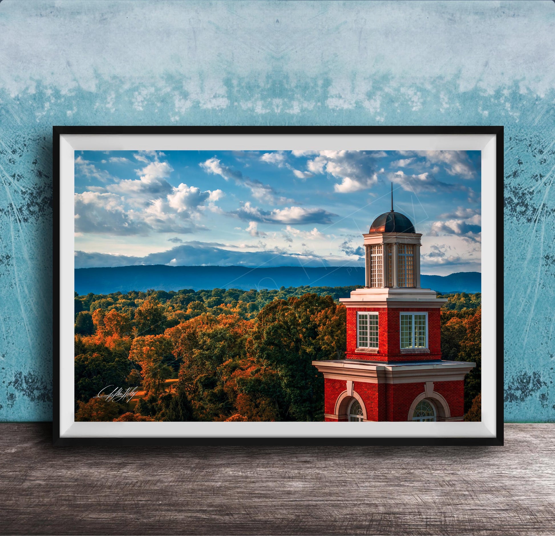 A framed photograph of a red brick building with a dome, set against a backdrop of a blue sky with clouds and a mountainous landscape.