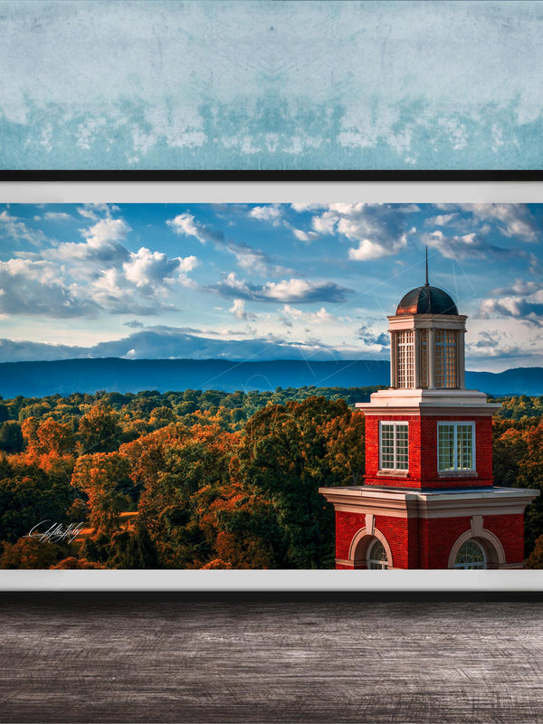 A framed photograph of a red brick building with a dome, set against a backdrop of a blue sky with clouds and a mountainous landscape.