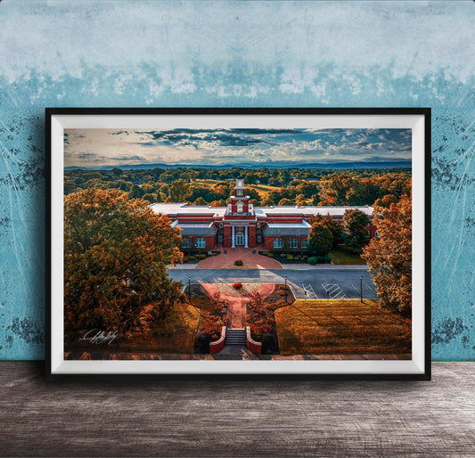 A framed aerial view of a red brick building with a clock tower, surrounded by trees with autumn foliage, set against a blue sky with clouds.
