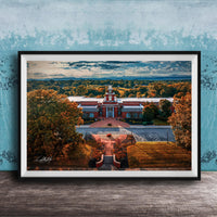 A framed aerial view of a red brick building with a clock tower, surrounded by trees with autumn foliage, set against a blue sky with clouds.