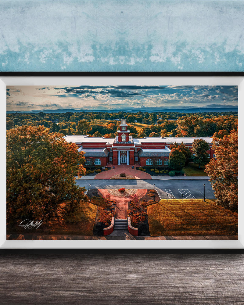 A framed aerial view of a red brick building with a clock tower, surrounded by trees with autumn foliage, set against a blue sky with clouds.