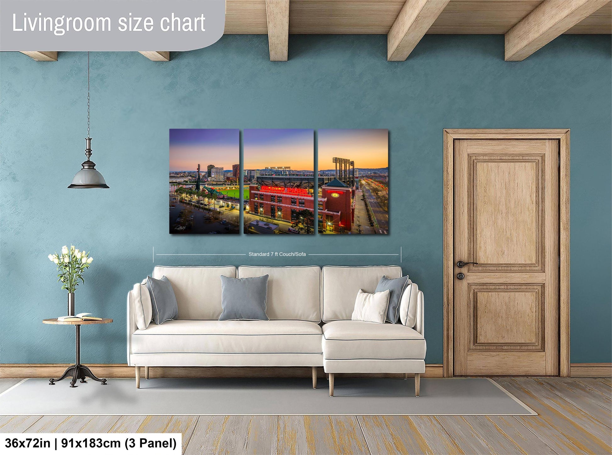 The image is a living room with a large wall art piece depicting a city skyline at sunset. The art piece is a collage of three separate images, each showing a different part of the cityscape. The living room has a white sofa and a wooden door, and the wall art is approximately 36 inches wide and 91 inches tall.
