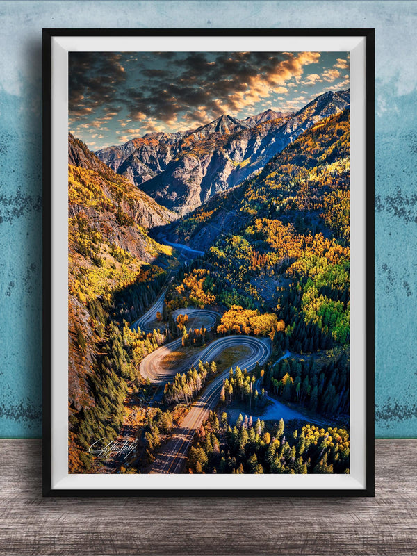 The image is a framed photograph of a winding mountain road surrounded by lush forests and autumnal trees, with a dramatic sky in the background.