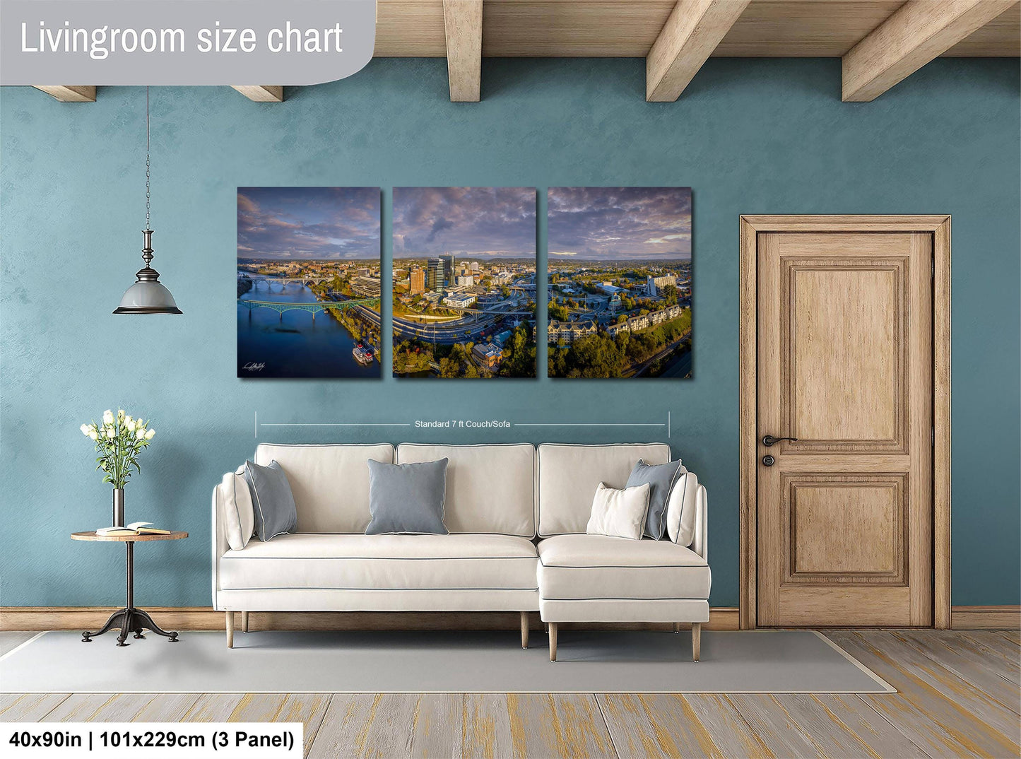 The image is a living room with a large white sofa, a wooden door, and a hanging light fixture. Above the sofa, there is a triptych of landscape photographs depicting a cityscape and a river. The wall behind the sofa has a living room size chart indicating the dimensions of the sofa and the triptych.