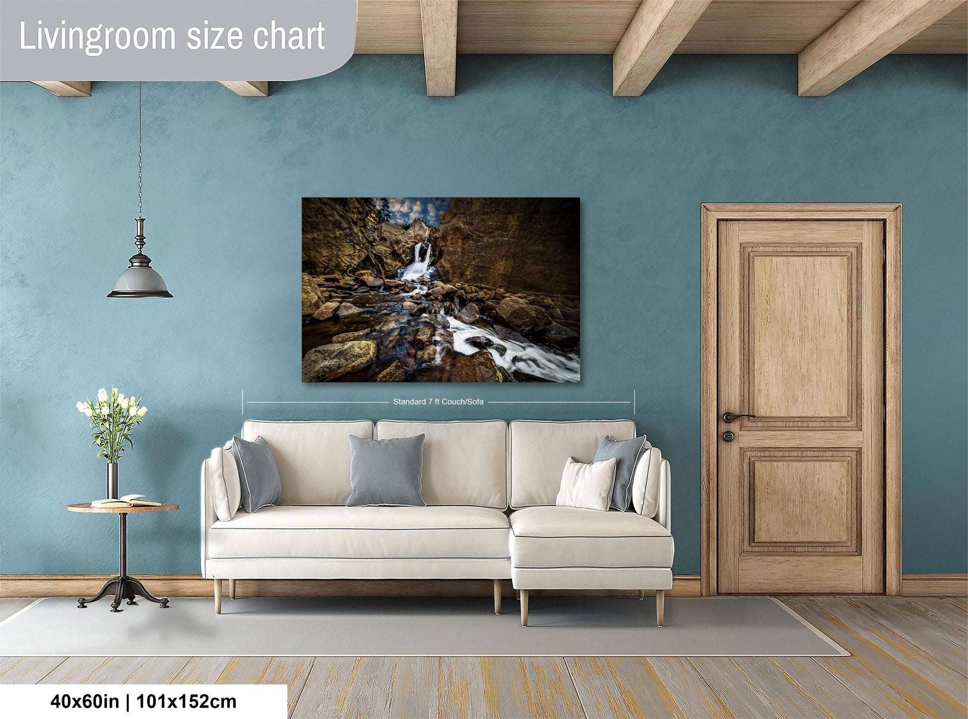 a living room with a large painting of a waterfall hanging on the wall. The painting is framed in a white frame and measures 40 inches by 101.5 centimeters. The room has a white sofa, a wooden door, and a small table with a vase of flowers.