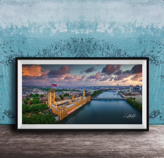 The image is a framed photograph of the Palace of Westminster, also known as the Houses of Parliament, in London, England. The photograph captures the iconic skyline of the city, with the River Thames flowing in the foreground. The sky is filled with vibrant colors, suggesting a beautiful sunset or sunrise. The photograph is framed and displayed on a wooden surface, creating a visually appealing and artistic composition.