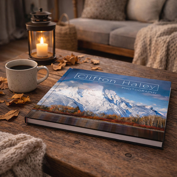 a wooden coffee table with a book titled "Clifton Haley Photography" on it, along with a lit candle, a mug of coffee, and some fallen leaves.