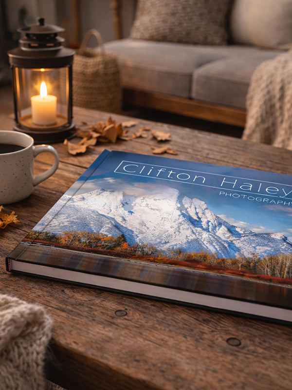 a wooden coffee table with a book titled "Clifton Haley Photography" on it, along with a lit candle, a mug of coffee, and some fallen leaves.