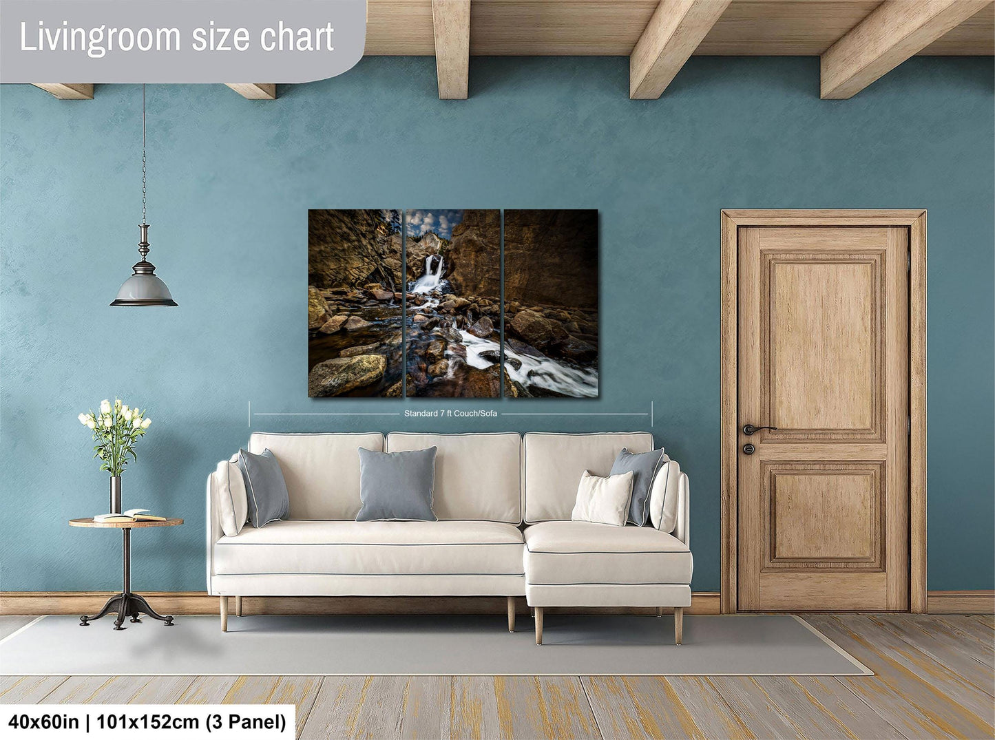 The image is a living room with a large painting of a waterfall hanging on the wall. The painting is framed and measures 40 inches by 101 inches, with a 3-panel layout. The room has a white sofa, a wooden door, and a small table with a vase of flowers.