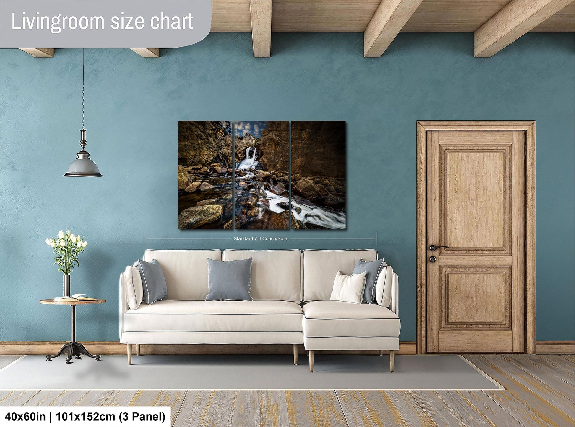 The image is a living room with a large painting of a waterfall hanging on the wall. The painting is framed and measures 40 inches by 101 inches, with a 3-panel layout. The room has a white sofa, a wooden door, and a small table with a vase of flowers.