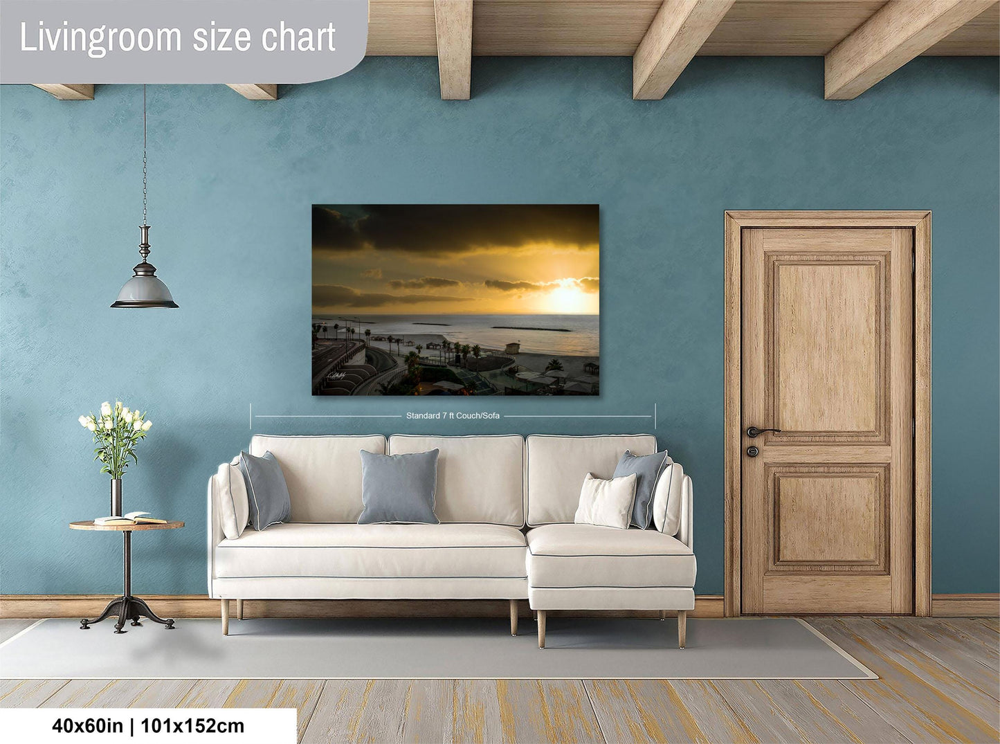 a living room with a large painting of a sunset over the ocean hanging on the wall. The painting is framed and measures 40 inches by 101.5 centimeters. The room has a white sofa, a wooden door, and a small table with a vase of flowers.