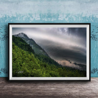 The image is a framed photograph of a mountainous landscape with a forested area and a cloudy sky.