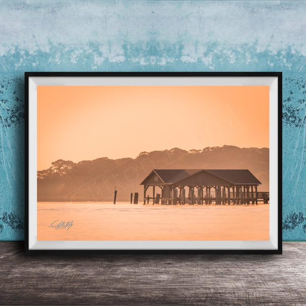 A framed photograph of a wooden pier extending into a body of water, with a warm orange and yellow sunset sky in the background.