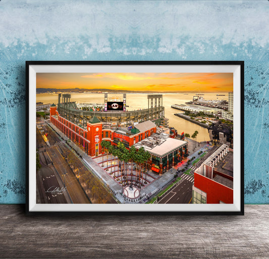 The image is a framed aerial view of a cityscape with a large stadium, buildings, and a body of water in the background.