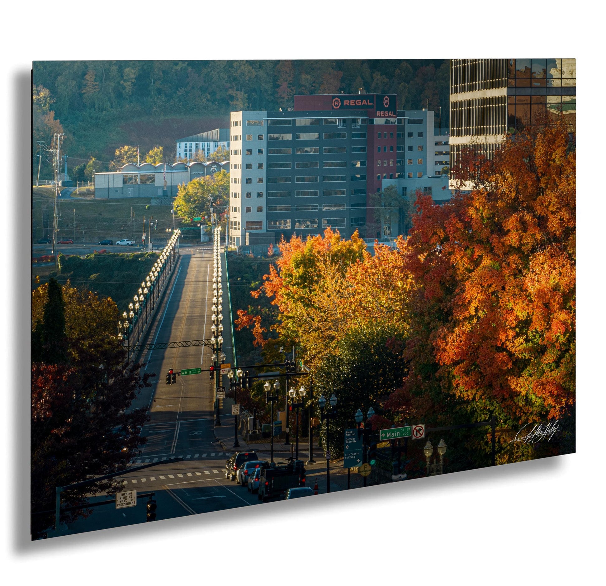 a cityscape with a large building, a bridge, and trees with autumn foliage. The sky is clear and blue, and the sun is shining brightly, creating a warm and vibrant atmosphere.
