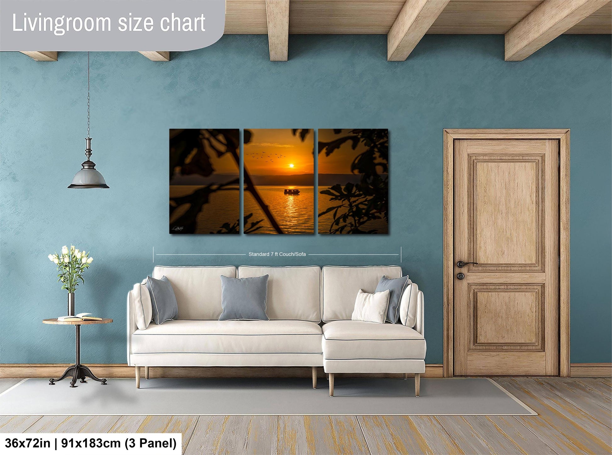 The image is a living room with a large, three-panel wall art depicting a sunset over the ocean. The wall art is hanging above a white sofa, and there is a wooden door and a small table with a vase of flowers nearby. The living room has a blue wall and a wooden floor.