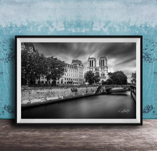 A black and white photograph of the Notre-Dame Cathedral in Paris, France, with a river and trees in the foreground.