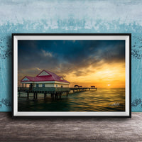 A framed photograph depicting a serene pier scene at sunset, with a red-roofed building and a wooden pier extending into the calm waters.