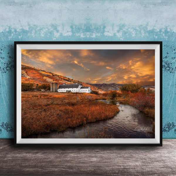 a picturesque rural landscape with a farmhouse nestled among rolling hills and a winding stream. The sky is filled with vibrant orange and yellow hues, creating a warm and serene atmosphere. The overall scene is a beautiful representation of nature and the tranquility of rural life.