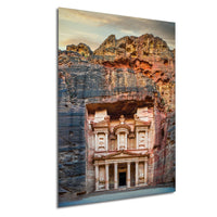 a large, ancient stone structure carved into a cliff face, with a carved facade and columns. The structure appears to be a carved temple or monument, set against a backdrop of rocky cliffs and a cloudy sky.