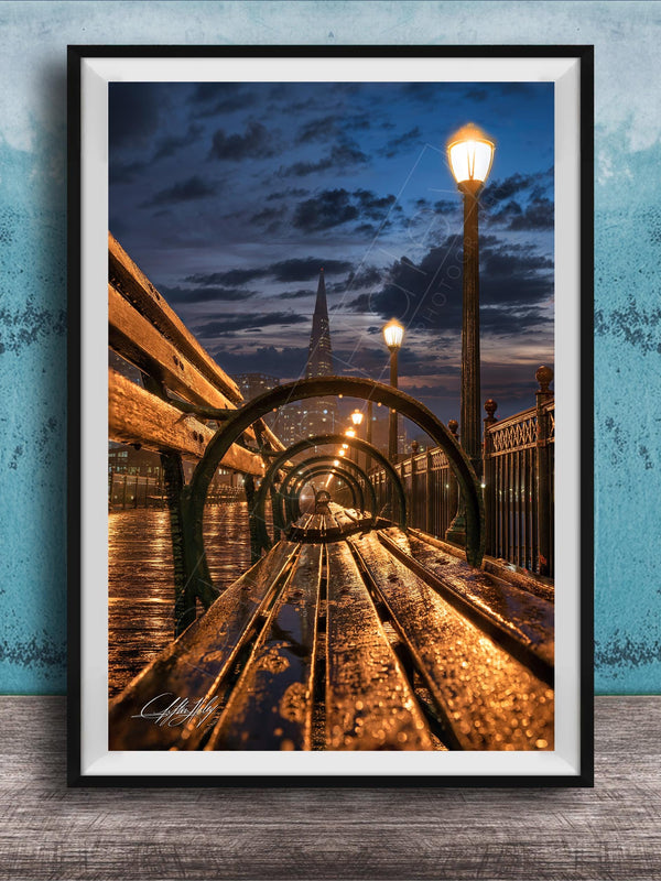 A wooden bench on a bridge at night, with a city skyline and street lamps in the background, creating a serene and atmospheric scene.