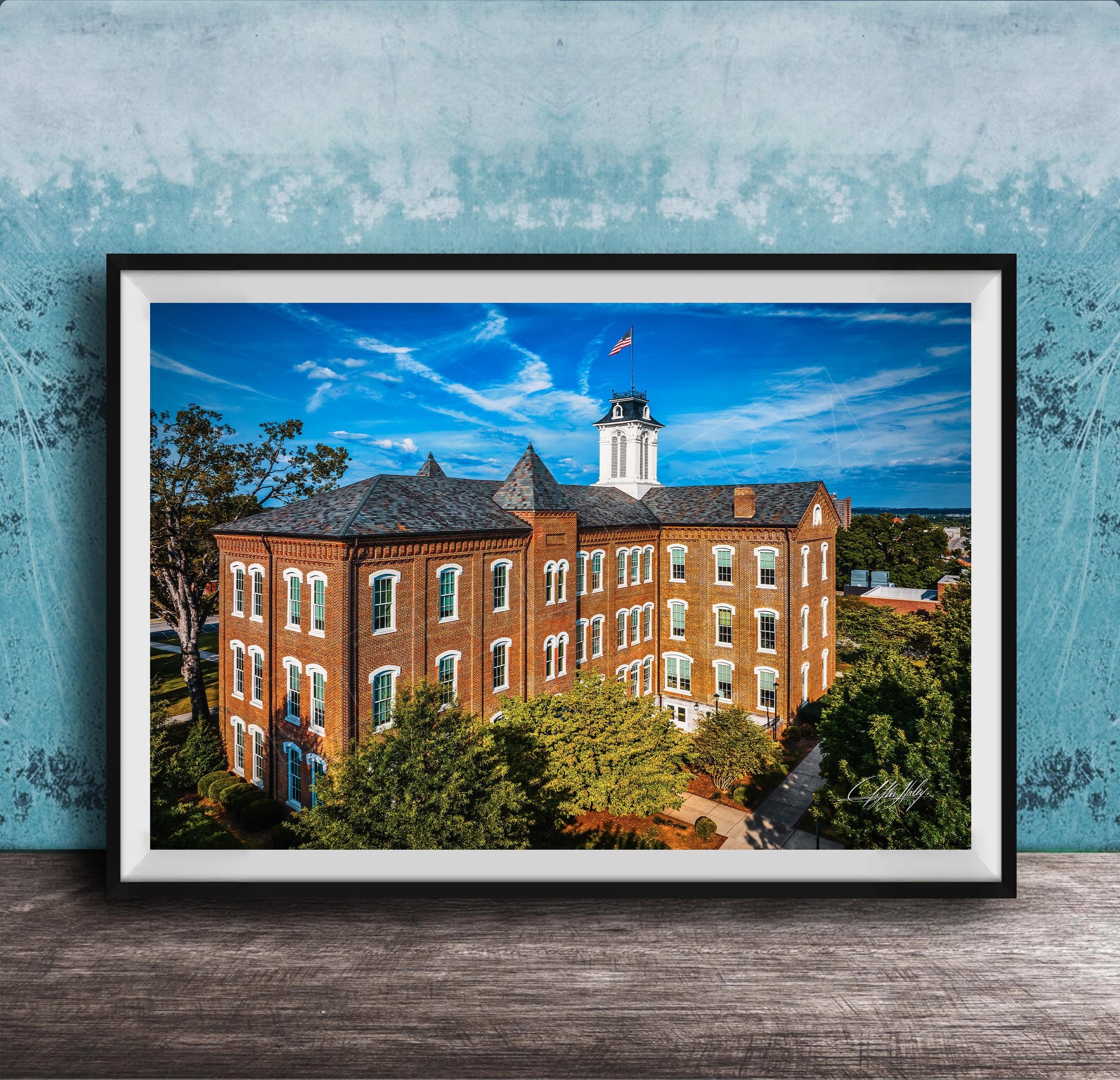 A framed photograph of a large brick building with a clock tower, surrounded by trees and a blue sky with clouds.