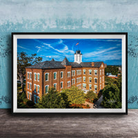 A framed photograph of a large brick building with a clock tower, surrounded by trees and a blue sky with clouds.