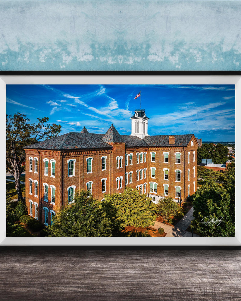 A framed photograph of a large brick building with a clock tower, surrounded by trees and a blue sky with clouds.