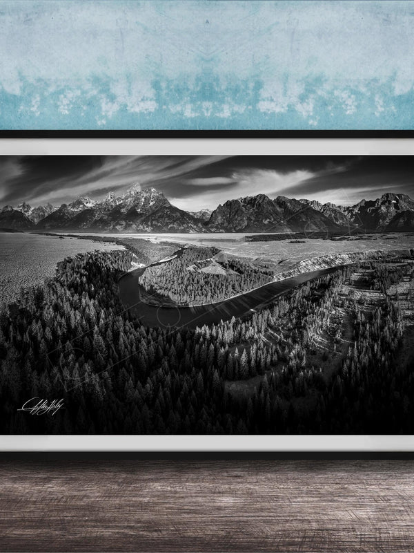 A black and white landscape photograph depicting a mountainous, forested area with a winding river or stream in the foreground.