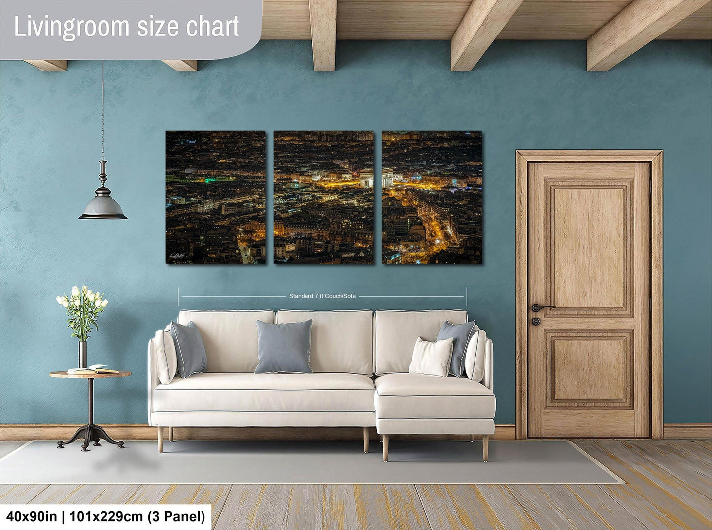 The image is a living room with a large, three-panel wall art depicting a cityscape at night. The wall art is mounted above a white sofa, and there is a wooden door and a small table with a vase of flowers nearby. The living room has a blue wall and wooden flooring.