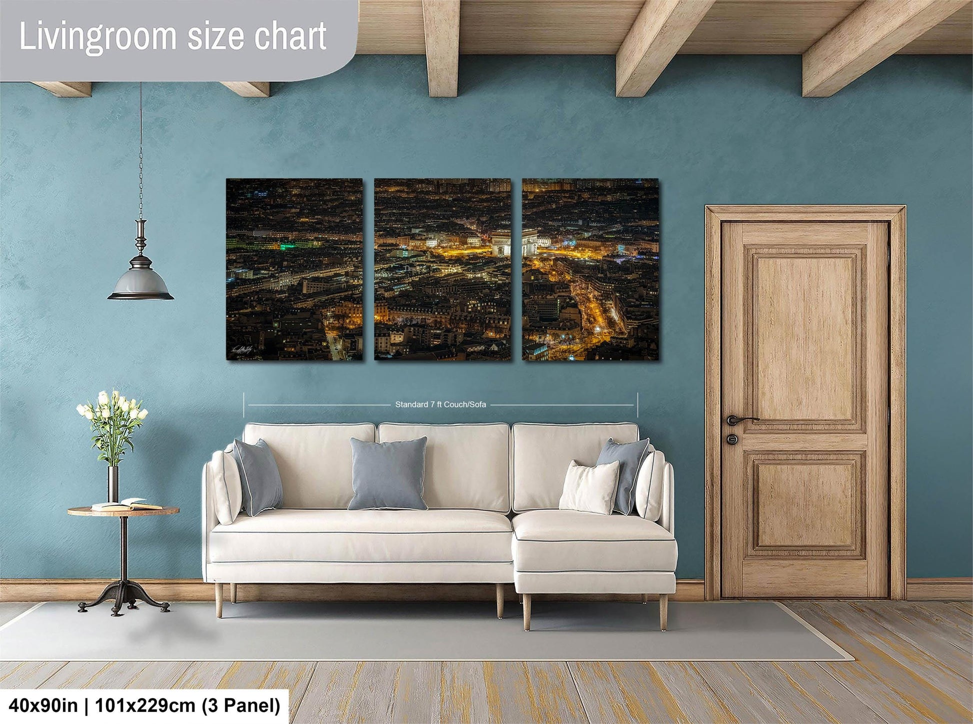 The image is a living room with a large, three-panel wall art depicting a cityscape at night. The wall art is mounted above a white sofa, and there is a wooden door and a small table with a vase of flowers nearby. The living room has a blue wall and wooden flooring.