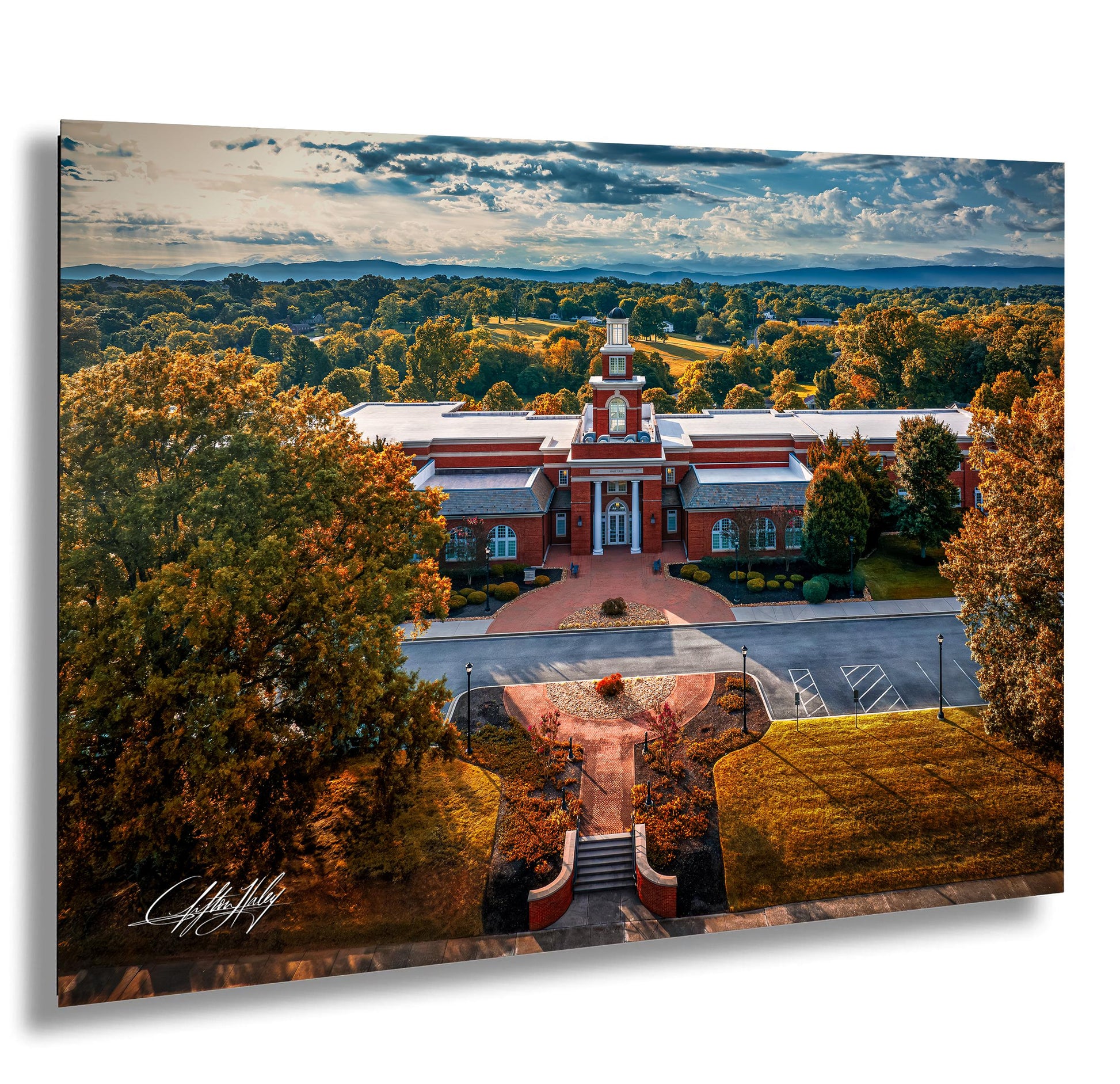 a large, red brick building with a prominent clock tower, surrounded by a lush, tree-lined landscape with a clear blue sky.