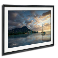 A framed photograph depicting a serene coastal scene with a sailboat on the water, a mountain range in the background, and a dramatic sky with clouds at sunset.